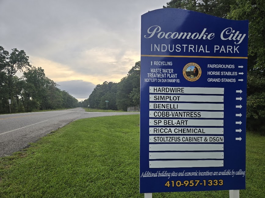 Pocomoke Industrial Park - Pocomoke City Arrowhead Blue Pocomoke City Industrial Park sign listing tenants and contact number 410-957-1333 beside a rural road with grass and trees on the sides.