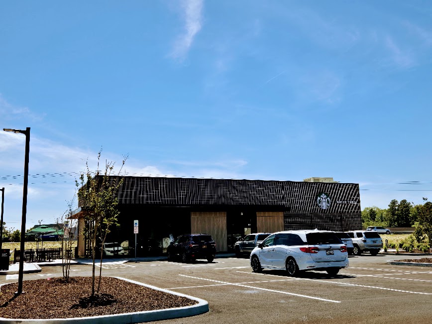 Starbucks photo - Pocomoke City Arrowhead Starbucks drive-thru storefront with a dark exterior, several cars parked in front on a sunny day under a blue sky.